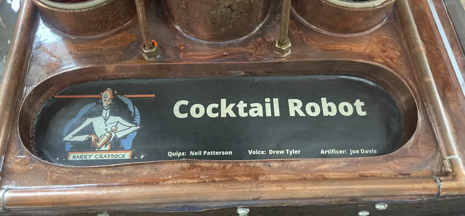 The maker's plate on the front of the machine: an illustration of Harry Craddock beside the words Cocktail Robot, and the credits Quips: Neil Patterson, Voice: Drew Tyler, Artificer: Joe Davis